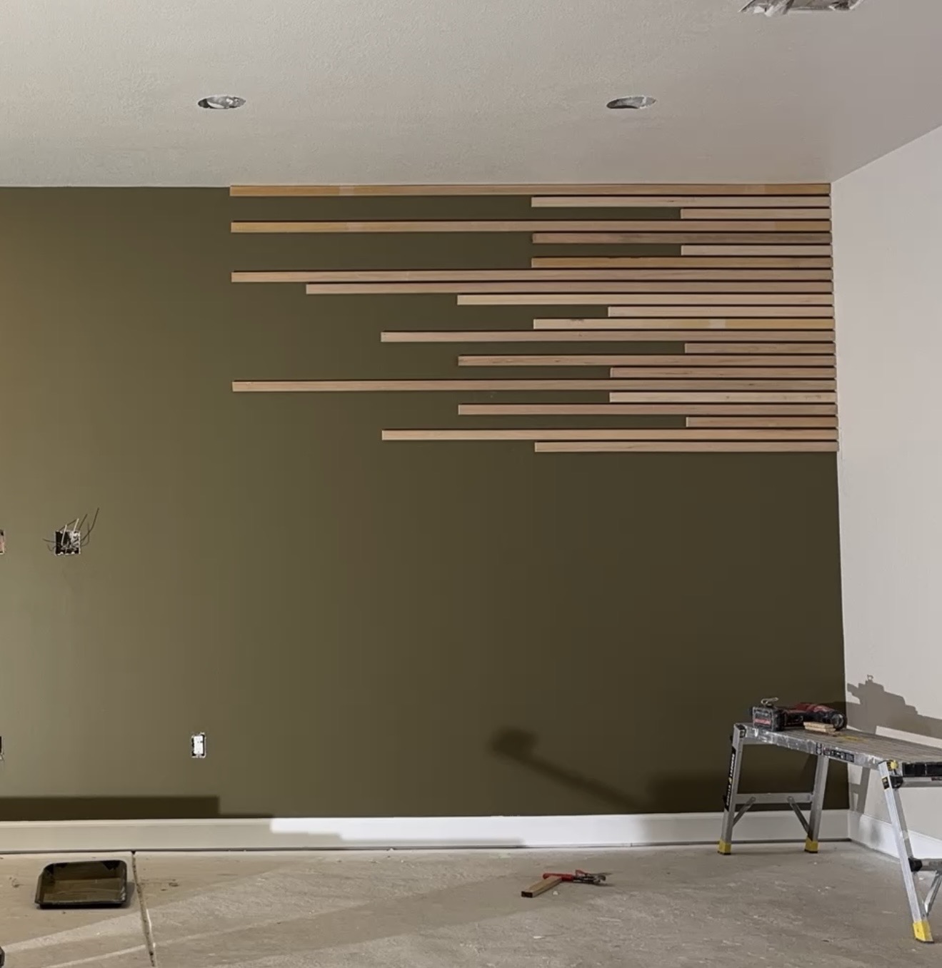 Olive-green wall with light wood slat strips being installed in a staggered pattern, sawhorse and tools nearby